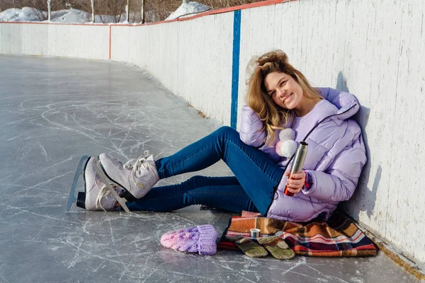 Adorável jovem relaxante depois de andar de patins no gelo e beber bebida quente de termo pot na pista de gelo . — Fotografia de Stock