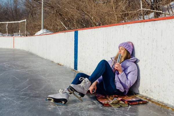 Krásná mladá žena relaxační po jízdě na bruslích a pití teplého nápoje z termo hrnce na kluzišti. — Stock fotografie