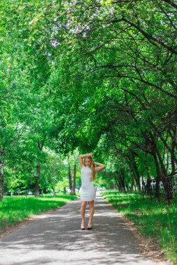 Ağaçların altında duran, güzel beyaz elbiseler giyen çekici sarışın bir kadının portresi..