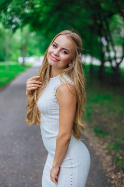 Ağaçların altında duran, güzel beyaz elbiseler giyen çekici sarışın bir kadının portresi..