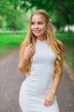 Ağaçların altında duran, güzel beyaz elbiseler giyen çekici sarışın bir kadının portresi..