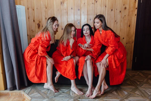 Four beautiful young women dressed in red bathrobe sitting on a couch and waching mobile phone — Stock Photo, Image