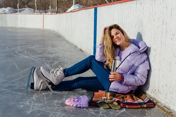 Krásná mladá žena relaxační po jízdě na bruslích a pití teplého nápoje z termo hrnce na kluzišti. — Stock fotografie