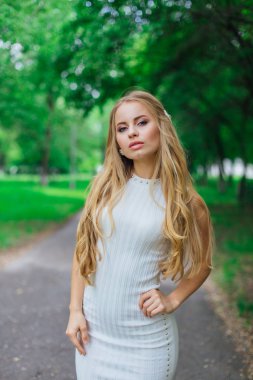 Ağaçların altında duran, güzel beyaz elbiseler giyen çekici sarışın bir kadının portresi..