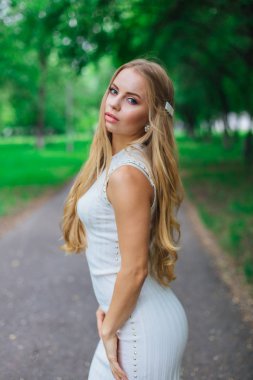 Ağaçların altında duran, güzel beyaz elbiseler giyen çekici sarışın bir kadının portresi..