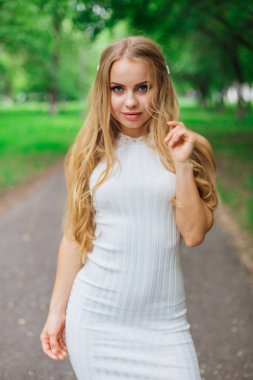 Ağaçların altında duran, güzel beyaz elbiseler giyen çekici sarışın bir kadının portresi..