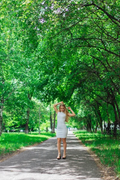 Porträtt av en charmig blond kvinna klädd i vacker vit klänning stående på vägen under träd. — Stockfoto