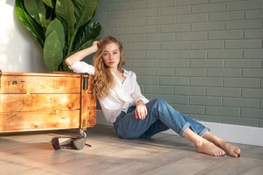 Genç ve güzel bir kadın çıplak ayakla yerde oturuyor, tahta kutu ve yeşil bitkinin yanında.