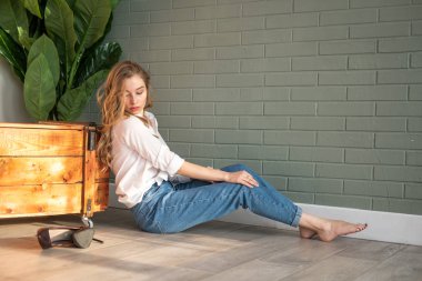 Genç ve güzel bir kadın çıplak ayakla yerde oturuyor, tahta kutu ve yeşil bitkinin yanında.