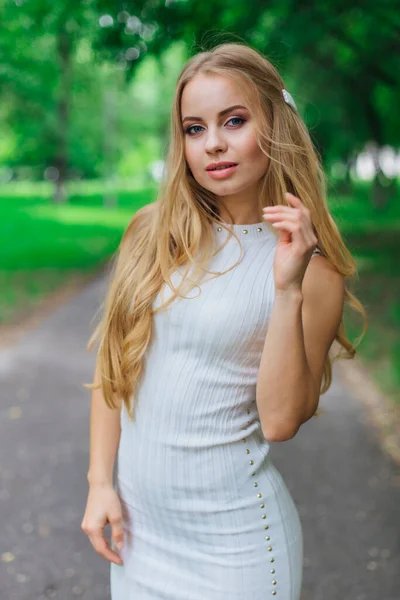 Retrato de una encantadora mujer rubia con hermoso vestido blanco de pie en la carretera bajo los árboles . — Foto de Stock