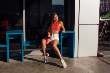 Beyaz şort ve spor ayakkabı giyen şık mutlu genç esmer kadın gitmek için kahve pembe fincan tutan.