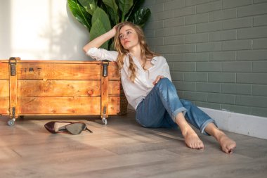 Genç ve güzel bir kadın çıplak ayakla yerde oturuyor, tahta kutu ve yeşil bitkinin yanında.