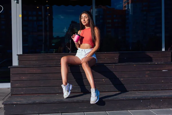 Elegante mujer morena feliz joven con pantalones cortos blancos y zapatillas de deporte con taza de café rosa para ir sentado al lado de la cafetería . —  Fotos de Stock