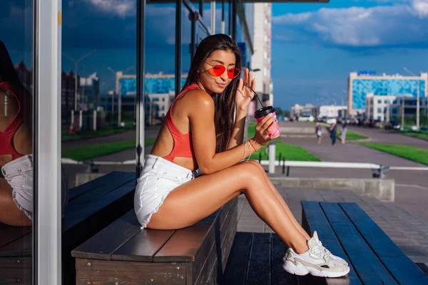 Elegante feliz jovem morena mulher vestindo branco shorts e tênis segurando rosa xícara de café para ir sentado ao lado de café . — Fotografia de Stock