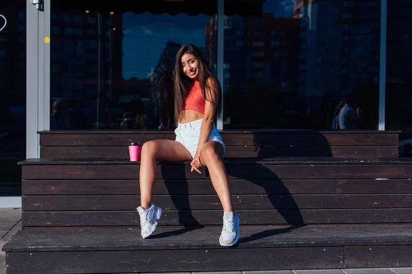 Elegante feliz jovem morena mulher vestindo branco shorts e tênis segurando rosa xícara de café para ir sentado ao lado de café . — Fotografia de Stock