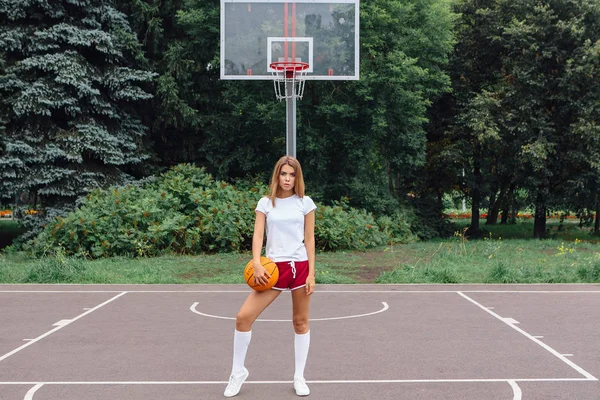 Menina bonita vestida com t-shirt branca, shorts e tênis, joga com uma bola em um campo de basquete . — Fotografia de Stock