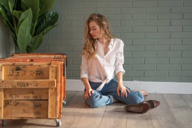 Genç ve güzel bir kadın çıplak ayakla yerde oturuyor, tahta kutu ve yeşil bitkinin yanında.