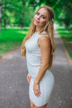 Ağaçların altında duran, güzel beyaz elbiseler giyen çekici sarışın bir kadının portresi..