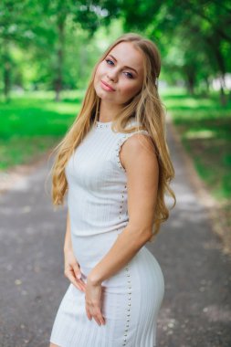 Ağaçların altında duran, güzel beyaz elbiseler giyen çekici sarışın bir kadının portresi..