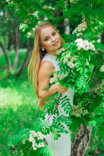 一个迷人的金发女人穿着漂亮的白色连衣裙站在一棵开着白花的树旁边的画像. — 图库照片