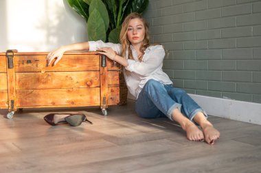 Genç ve güzel bir kadın çıplak ayakla yerde oturuyor, tahta kutu ve yeşil bitkinin yanında.