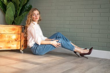 Genç ve güzel bir kadın yerde, tahta kutunun ve yeşil bitkinin yanında oturuyor.