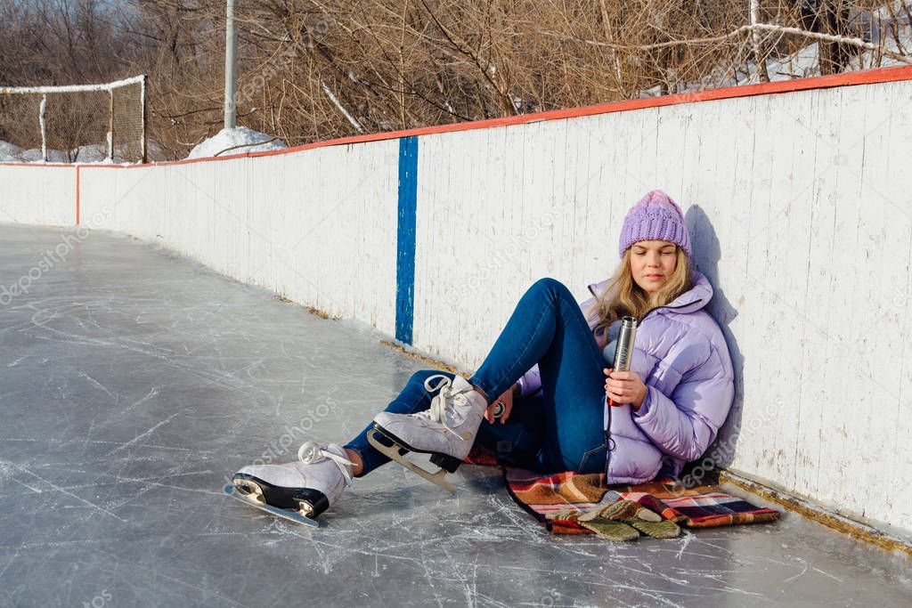 Lovely young woman relaxing after riding ice skates and drinking hot drink from termo pot on the ice rink.