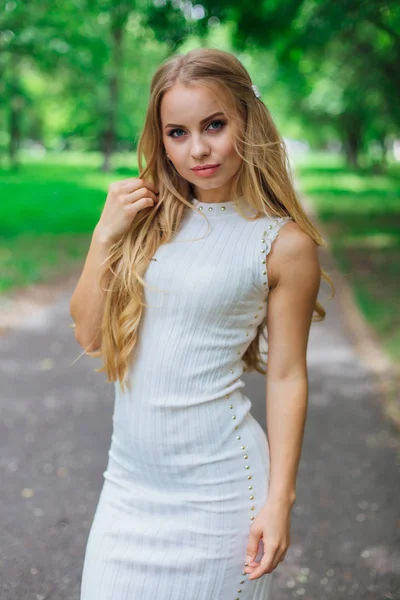 Portrait of a charming blond woman wearing beautiful white dress standing on the road under trees. — Stock Photo, Image