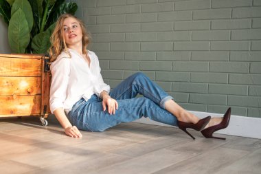 Genç ve güzel bir kadın yerde, tahta kutunun ve yeşil bitkinin yanında oturuyor.