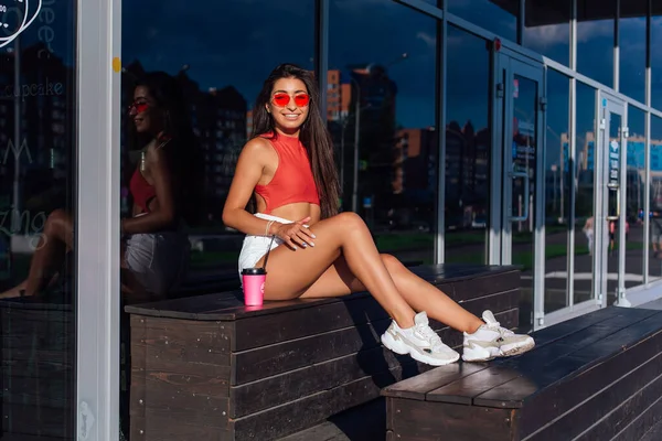 Elegante feliz jovem morena mulher vestindo branco shorts e tênis segurando rosa xícara de café para ir sentado ao lado de café . — Fotografia de Stock