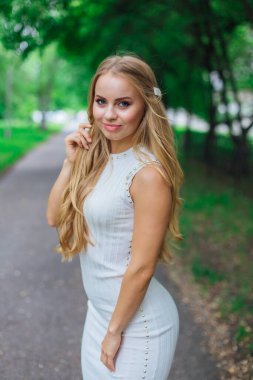 Ağaçların altında duran, güzel beyaz elbiseler giyen çekici sarışın bir kadının portresi..