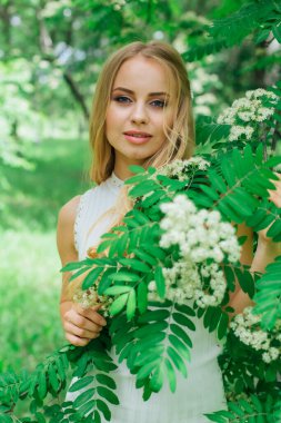 Beyaz çiçekli Rowan ağacının yanında duran güzel, beyaz elbiseli sarışın bir kadının portresi..