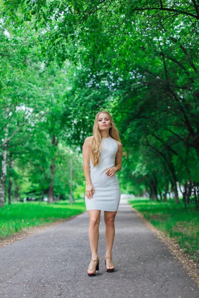 Портрет чарівної блондинки в красивій білій сукні, що стоїть на дорозі під деревами . — стокове фото