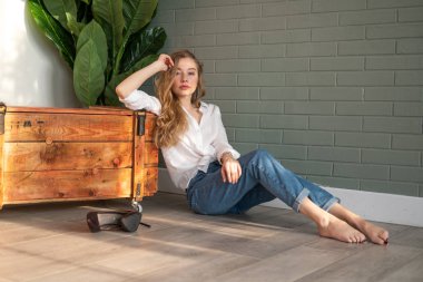 Genç ve güzel bir kadın çıplak ayakla yerde oturuyor, tahta kutu ve yeşil bitkinin yanında.