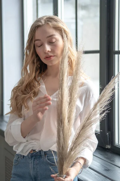 Giovane bella donna bionda in piedi accanto alla finestra con vaso con piante secche — Foto Stock