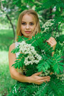 Güzel beyaz elbiseler giyen çekici bir sarışının bahar portresi çiçek açan kürek ağacının yanında beyaz çiçeklerle duruyor..