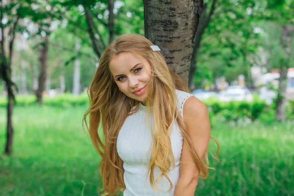 一位漂亮的金发女子穿着漂亮的白色连衣裙 站在公园里一棵树旁的春日画像 — 图库照片
