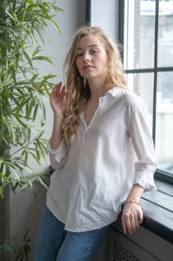 Pencerenin yanında beyaz gömlek ve kot pantolon giyen güzel sarışın genç bir kadın var. Arka planda yeşil bitkiler ve penceresi olan bir kızın sabah portresi.