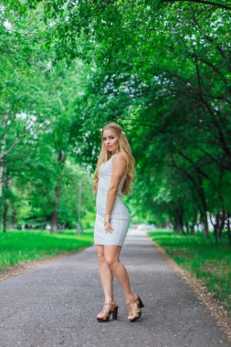 Yeşil ağaçların altında duran yolda duran güzel beyaz elbiseli çekici sarışın bir kadının bahar portresi..