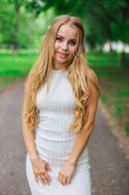 Yeşil ağaçların altında duran yolda duran güzel beyaz elbiseli çekici sarışın bir kadının bahar portresi..