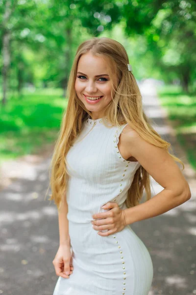 Yeşil ağaçların altında duran yolda duran güzel beyaz elbiseli çekici sarışın bir kadının bahar portresi..