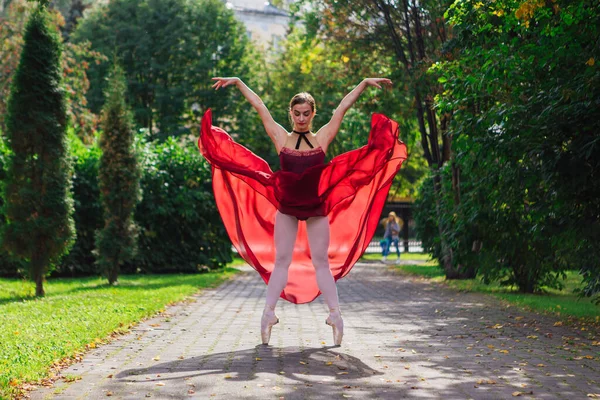 Woman ballerina in red ballet dress dancing in pointe shoes in autumn park. Ballerina standing in beautiful ballet pose