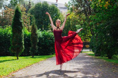 Sonbahar parkında kırmızı bale elbiseli kadın balerin, sivri uçlu ayakkabılarla dans ediyor. Balerin güzel bir bale pozisyonunda duruyor.