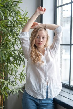 Pencerenin yanında beyaz gömlek ve kot pantolon giyen güzel sarışın genç bir kadın var. Arka planda yeşil bitkiler ve penceresi olan bir kızın sabah portresi.