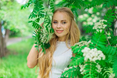 Güzel beyaz elbiseler giyen çekici bir sarışının bahar portresi çiçek açan kürek ağacının yanında beyaz çiçeklerle duruyor..