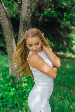 Güzel beyaz elbiseler giyen çekici bir sarışının bahar portresi. Parktaki Rowan ağacının yanında çimlerin üzerinde duruyor..