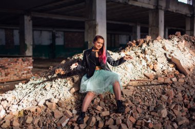 Pembe saçlı genç bir kız çöken bir binada, tuğlaların üzerinde oturuyor.