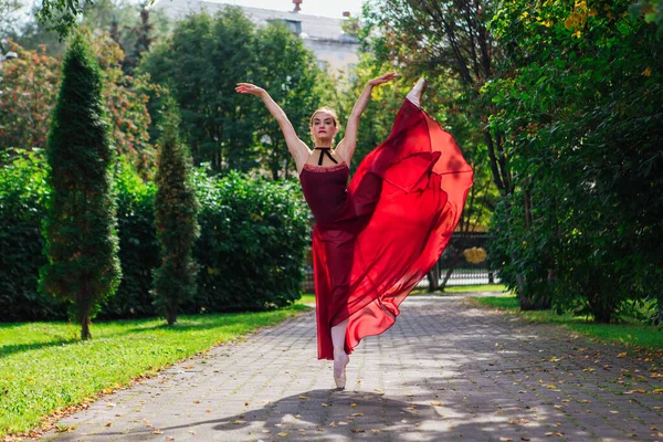 Woman ballerina in red ballet dress dancing in pointe shoes in autumn park. Ballerina standing in beautiful ballet pose