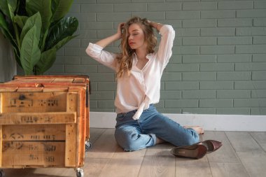 Genç, sarışın, beyaz gömlek ve kot pantolon giyen, yerde, tahta kutu ve yeşil bitkinin yanında çıplak ayakla oturan güzel bir kadın. Pencereden gün ışığında oturan bir kız. Boşluğu kopyala