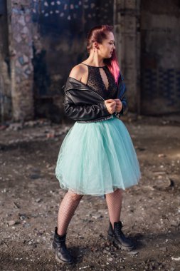 Deri ceketli ve mavi etekli pembe saçlı genç kız yıkılmış binanın içinde yıkıntılarla çevrili duruyor. Arka planda eski mozaik duvarın yanında duran kadın..